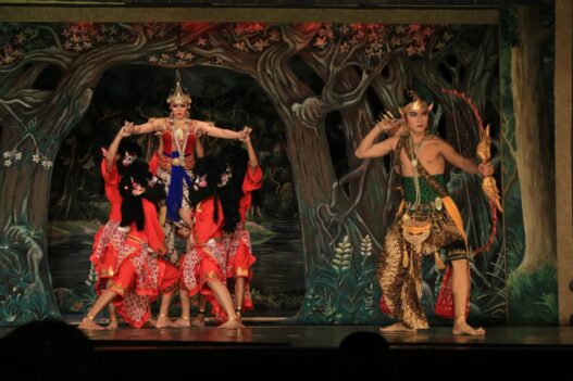 Penari wayang orang berbusana kesatria memegang busur panah berhadapan dengan tokoh putri dan raksasa di panggung teater.