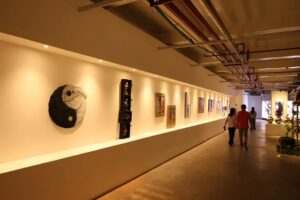 Pengunjung berjalan di lorong pameran seni rupa dengan sorotan lampu hangat yang menerangi deretan lukisan dan karya seni di dinding.