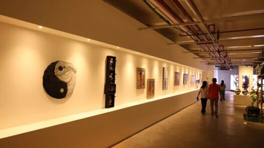 Pengunjung berjalan di lorong pameran seni rupa dengan sorotan lampu hangat yang menerangi deretan lukisan dan karya seni di dinding.
