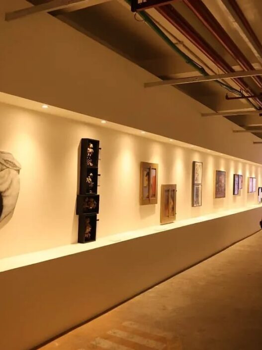 Pengunjung berjalan di lorong pameran seni rupa dengan sorotan lampu hangat yang menerangi deretan lukisan dan karya seni di dinding.