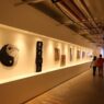 Pengunjung berjalan di lorong pameran seni rupa dengan sorotan lampu hangat yang menerangi deretan lukisan dan karya seni di dinding.
