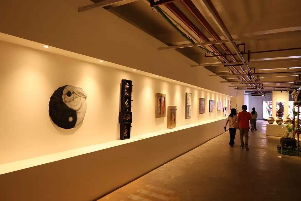 Pengunjung berjalan di lorong pameran seni rupa dengan sorotan lampu hangat yang menerangi deretan lukisan dan karya seni di dinding.