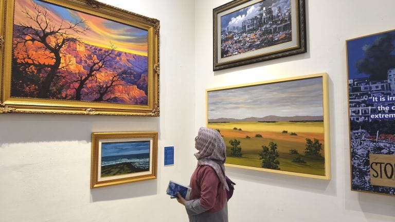 Seorang pengunjung berhijab sedang mengamati deretan lukisan lanskap alam yang terpajang di dinding galeri.