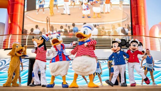 Karakter Disney dalam seragam pelaut meriah di panggung kapal pesiar Disney Cruise Line, berlatar belakang cerobong asap merah dan layar besar.