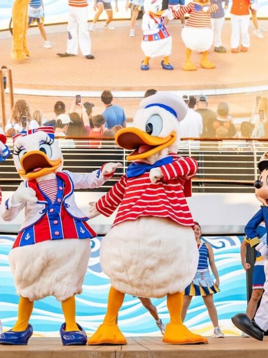 Karakter Disney dalam seragam pelaut meriah di panggung kapal pesiar Disney Cruise Line, berlatar belakang cerobong asap merah dan layar besar.