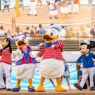 Karakter Disney dalam seragam pelaut meriah di panggung kapal pesiar Disney Cruise Line, berlatar belakang cerobong asap merah dan layar besar.