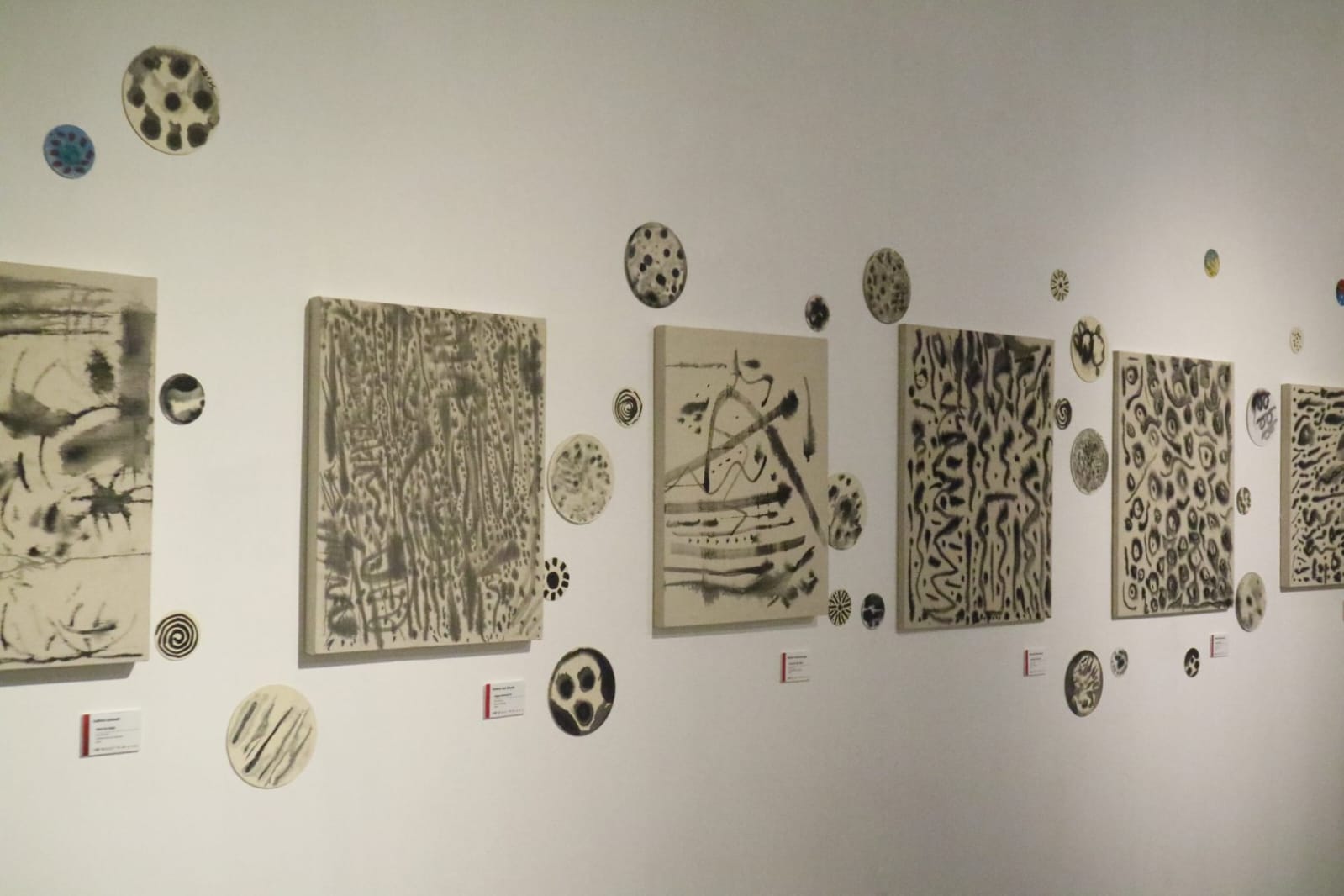 Deretan lukisan kanvas abstrak dan piringan keramik monokrom terpasang rapi di dinding putih galeri pameran.