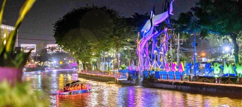 Perahu wisata melintas di Sungai Kalimas malam hari dengan latar patung hiu bercahaya dan landmark Taman Skate & BMX Surabaya.
