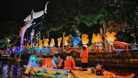 Wisatawan mengenakan pelampung oranye naik perahu di Sungai Kalimas Surabaya malam hari dengan latar patung hiu dan lampu hias SKATE & BMX.