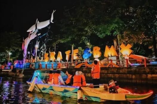 Wisatawan mengenakan pelampung oranye naik perahu di Sungai Kalimas Surabaya malam hari dengan latar patung hiu dan lampu hias SKATE & BMX.