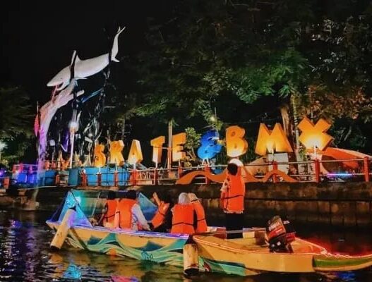 Wisatawan mengenakan pelampung oranye naik perahu di Sungai Kalimas Surabaya malam hari dengan latar patung hiu dan lampu hias SKATE & BMX.