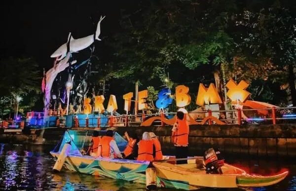 Wisatawan mengenakan pelampung oranye naik perahu di Sungai Kalimas Surabaya malam hari dengan latar patung hiu dan lampu hias SKATE & BMX.