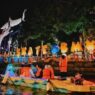 Wisatawan mengenakan pelampung oranye naik perahu di Sungai Kalimas Surabaya malam hari dengan latar patung hiu dan lampu hias SKATE & BMX.