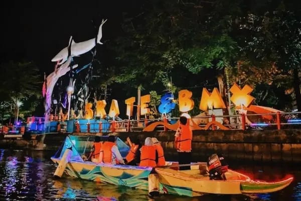 Wisatawan mengenakan pelampung oranye naik perahu di Sungai Kalimas Surabaya malam hari dengan latar patung hiu dan lampu hias SKATE & BMX.