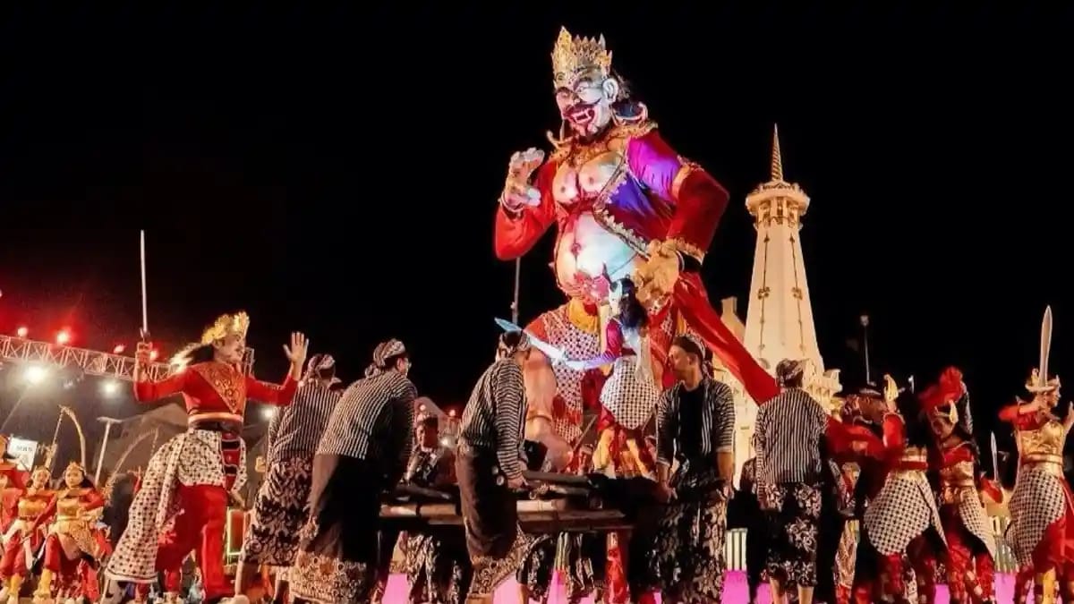 Figur raksasa wayang berbusana merah diusung para peserta karnaval di depan Tugu Yogyakarta saat malam hari.