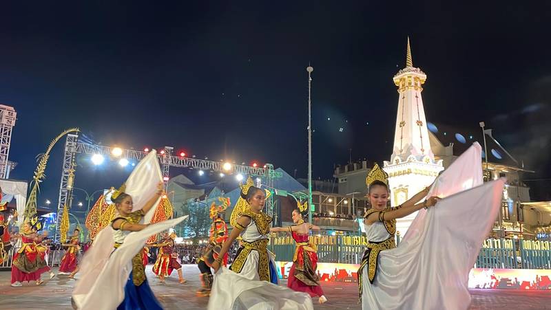 Para penari berbusana tradisional membentangkan kain putih saat tampil di jalan berlatar Tugu Yogyakarta yang bercahaya di malam hari.