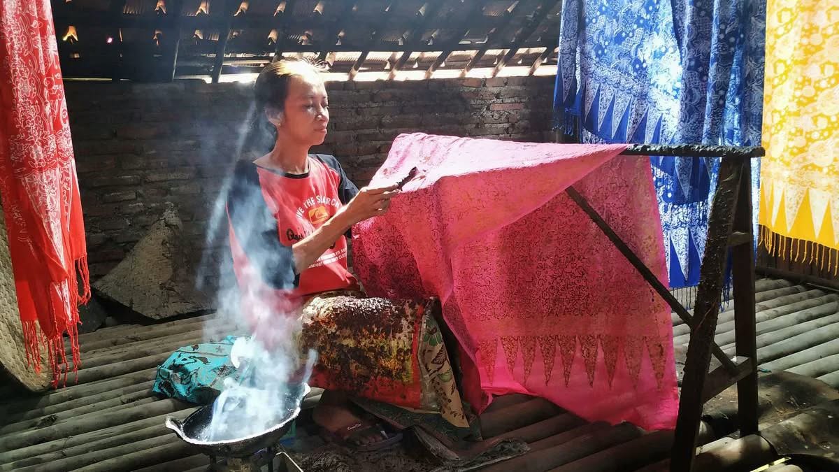 Seorang wanita pengrajin menorehkan lilin malam menggunakan canting pada kain batik warna merah muda di atas gawangan kayu.