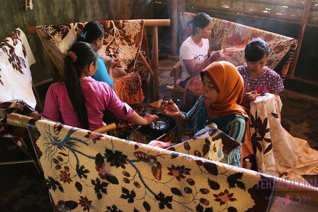 Sejumlah wanita duduk melingkar menggoreskan malam panas menggunakan canting pada kain batik tulis yang dibentangkan di gawangan kayu.