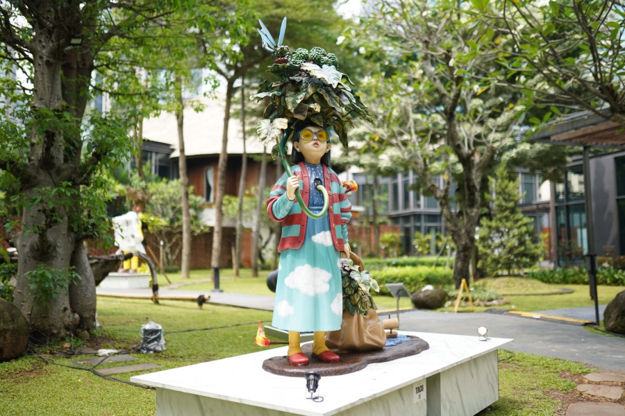Instalasi patung figur anak perempuan berbaju biru motif awan dan berkacamata kuning yang memegang payung dedaunan di taman terbuka.