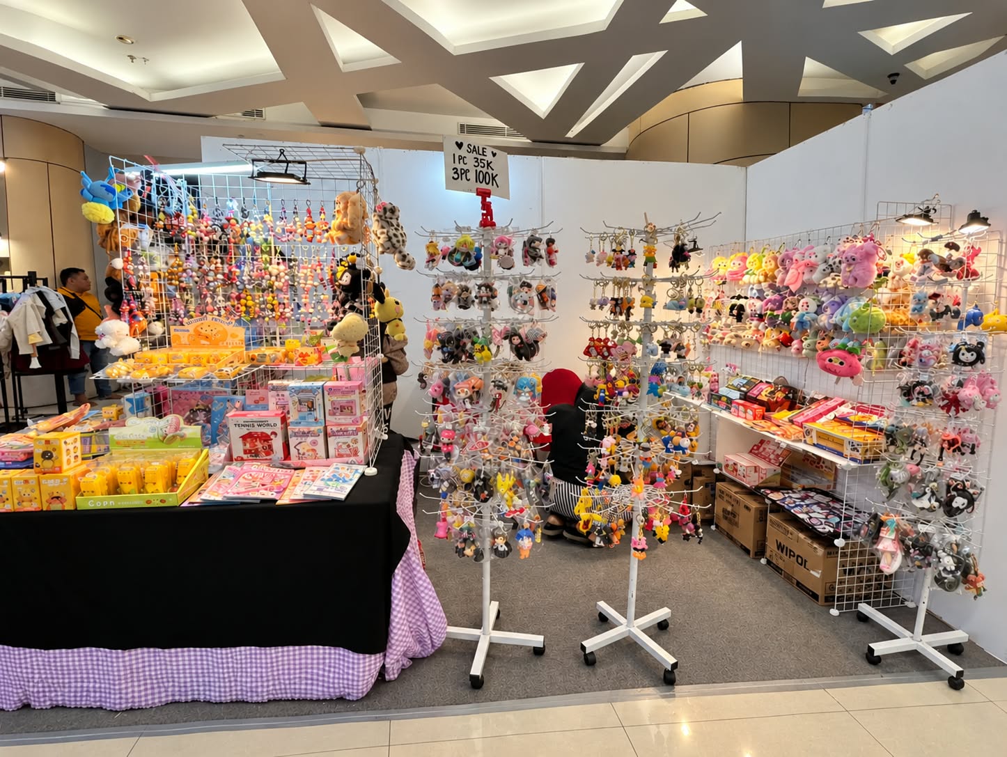 Stand bazar yang menjual aneka gantungan kunci karakter, boneka mini, dan mainan anak yang dipajang rapi pada rak putar serta dinding kisi-kisi.