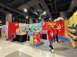 Booth pameran Komodo Food yang menampilkan produk camilan Garlic Chips dan Shrimp Chips dengan staf berkaos merah di area bazar.