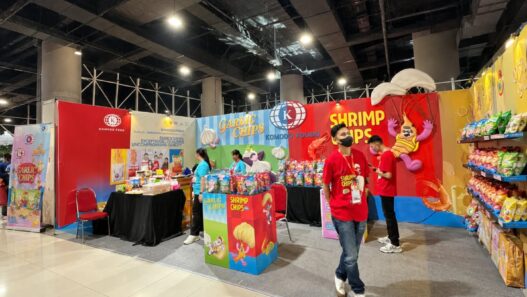 Booth pameran Komodo Food yang menampilkan produk camilan Garlic Chips dan Shrimp Chips dengan staf berkaos merah di area bazar.