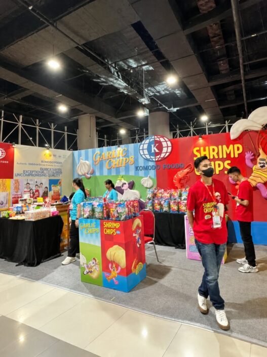 Booth pameran Komodo Food yang menampilkan produk camilan Garlic Chips dan Shrimp Chips dengan staf berkaos merah di area bazar.