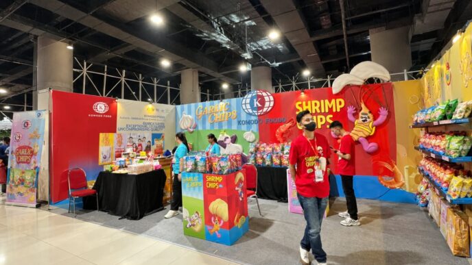 Booth pameran Komodo Food yang menampilkan produk camilan Garlic Chips dan Shrimp Chips dengan staf berkaos merah di area bazar.