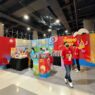 Booth pameran Komodo Food yang menampilkan produk camilan Garlic Chips dan Shrimp Chips dengan staf berkaos merah di area bazar.