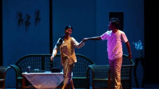 Pria dan wanita mengenakan pakaian tradisional-kasual bergandengan tangan dan tersenyum di atas panggung teater berlampu biru.