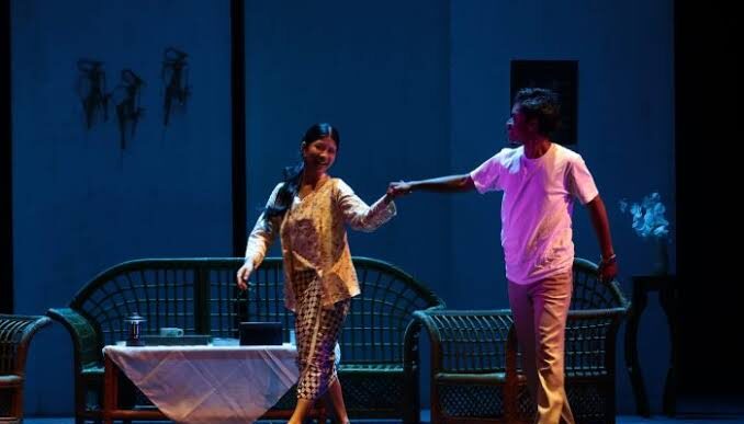 Pria dan wanita mengenakan pakaian tradisional-kasual bergandengan tangan dan tersenyum di atas panggung teater berlampu biru.