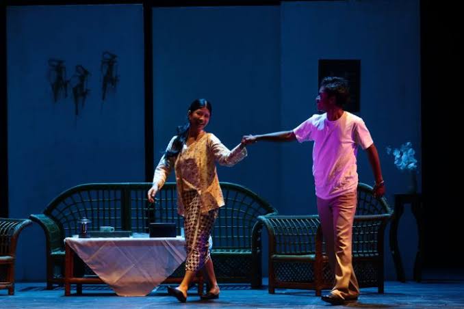 Pria dan wanita mengenakan pakaian tradisional-kasual bergandengan tangan dan tersenyum di atas panggung teater berlampu biru.