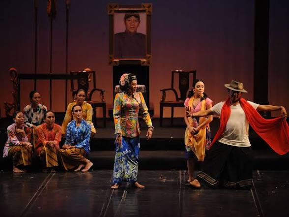Para pemain teater berkostum tradisional dan kain batik beraksi di panggung dengan latar lukisan potret besar.