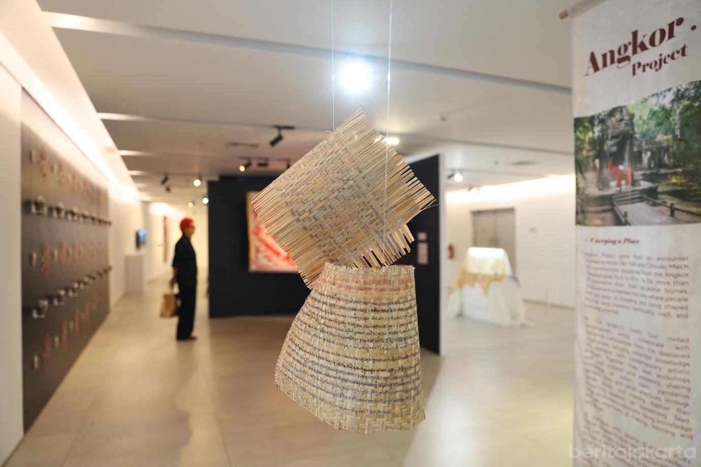 Anyaman bambu dan serat alam tergantung di tengah ruang galeri pameran dengan spanduk gantung Angkor Project di bagian kanan.