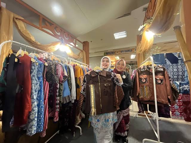 Dua wanita berhijab memegang dan memamerkan pakaian jaket motif batik di antara deretan gantungan busana fesyen lokal.