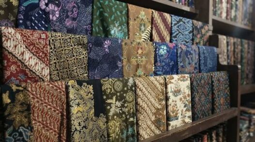 Berbagai lipatan kain batik dengan beragam motif dan warna tersusun rapi di rak kayu.