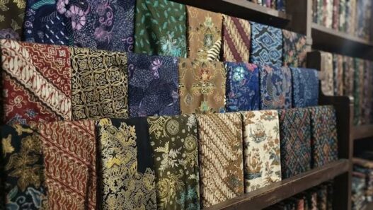 Berbagai lipatan kain batik dengan beragam motif dan warna tersusun rapi di rak kayu.
