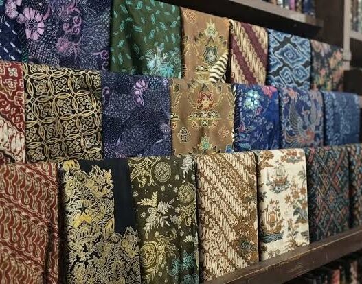 Berbagai lipatan kain batik dengan beragam motif dan warna tersusun rapi di rak kayu.
