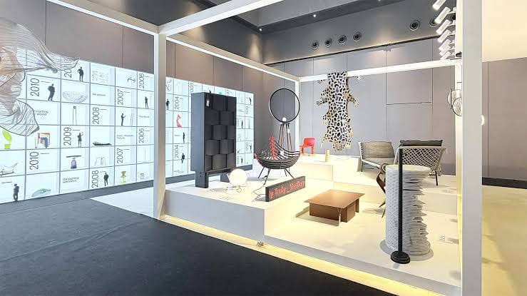 Sudut pameran desain menampilkan dinding linimasa berangka tahun, podium putih dengan berbagai furnitur modern, dan dekorasi kulit macan tutul di dinding.