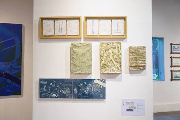 Dinding pameran putih menampilkan panel kertas bertekstur, bingkai kayu berisi lembaran kertas bernuansa organik, dan ilustrasi cetak biru di galeri seni.