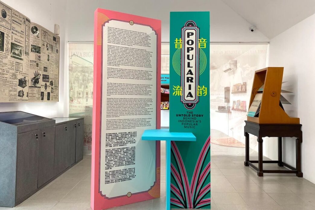 Panel informasi pameran berwarna merah muda dan hijau toska bertuliskan Popularia, dikelilingi arsip koran tua dan peralatan musik antik di ruang galeri.