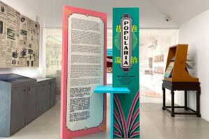 Panel informasi pameran berwarna merah muda dan hijau toska bertuliskan Popularia, dikelilingi arsip koran tua dan peralatan musik antik di ruang galeri.