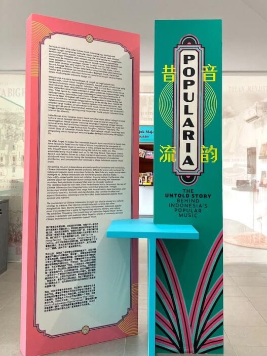 Panel informasi pameran berwarna merah muda dan hijau toska bertuliskan Popularia, dikelilingi arsip koran tua dan peralatan musik antik di ruang galeri.