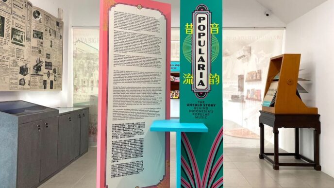 Panel informasi pameran berwarna merah muda dan hijau toska bertuliskan Popularia, dikelilingi arsip koran tua dan peralatan musik antik di ruang galeri.