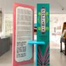 Panel informasi pameran berwarna merah muda dan hijau toska bertuliskan Popularia, dikelilingi arsip koran tua dan peralatan musik antik di ruang galeri.