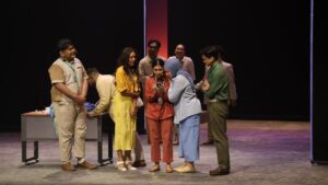 Para pemeran pertunjukan teater musikal Home Sweet Loan berkumpul di atas panggung dengan set ruang kantor, memperhatikan dua orang karakter yang sedang melihat layar ponsel bersama.