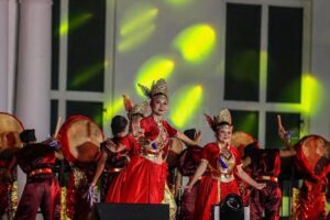 Para penari mengenakan busana adat merah emas dan hiasan kepala tradisional beraksi di panggung diiringi alat musik gendang besar serta sorotan lampu panggung yang dinamis.