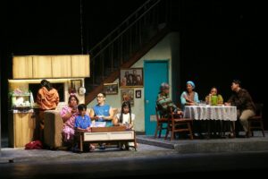 Para pemeran teater dalam set panggung berupa interior rumah modern dan area makan keluarga.
