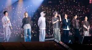 Tujuh anggota boy group K-Pop tampil bernyanyi di atas panggung konser besar dengan latar belakang lautan lightstick penonton yang menyala terang. 