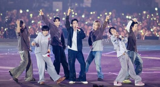 Enam anggota grup K-Pop ENHYPEN tampil energik di atas panggung konser besar dengan latar belakang penonton yang memegang lightstick menyala.